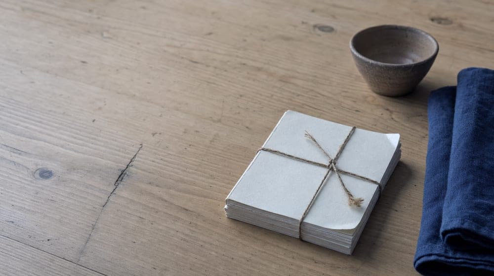 A short stack of blank paper bound with natural twine on a scrubbed pine table, beside a folded cloth and a small clay bowl, in flat overcast daylight.