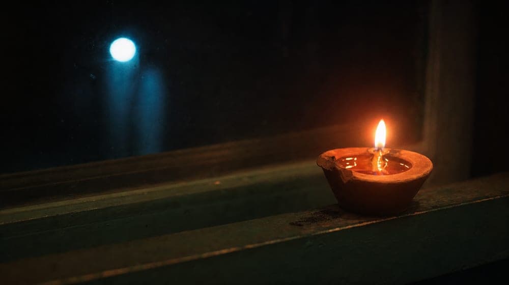 A small clay oil lamp burning alone on a dark windowsill at night, evoking care and the protection of something fragile.