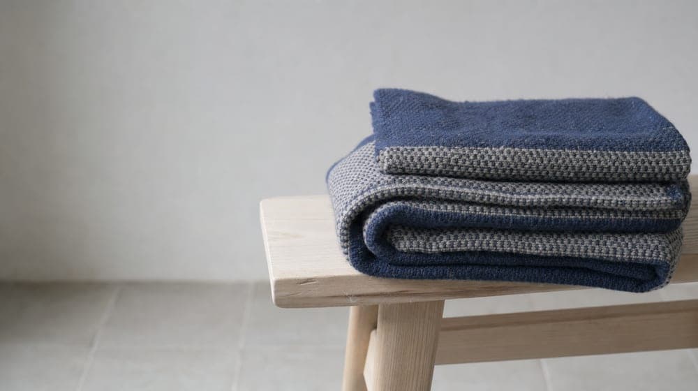 A handwoven wool blanket folded on a plain wooden bench in soft overcast light, a quiet space prepared for someone.